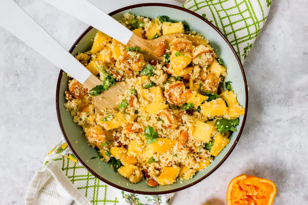 Grote kom met een salade van couscous, mango, scampi, amandelen, cashews, peterselie en basilicum met twee grote spatels erin
