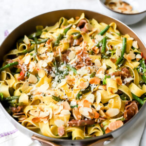 een grote pan met hoge randen waarin tagliatelle zit met boontjes, rode peper en parmaham. Afgewerkt met geroosterde amandelschilfers en parmezaan.