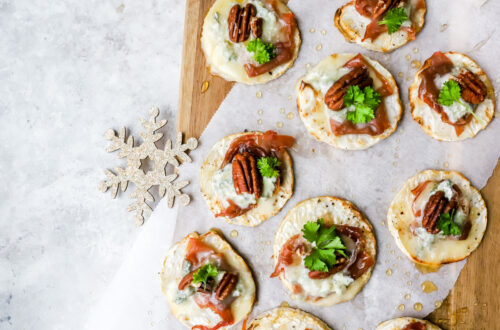 Bovenaanzicht van een plankje met toastjes van knolselder, parmaham, gorgonzola, pecannoten en peterselie en honing uit de oven.