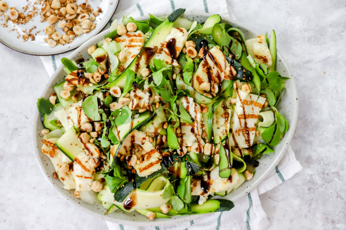 Grote schotel met een groene salade van komkommer, courgette en veldsla erop. Daarop ligt gegrilde halloumi, geroosterde noten, zaden en pitten. Afgewerkt met balsamicocreme.