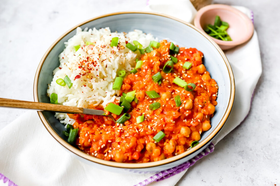 Een groot bord met rijst en rode curry van kikkererwten en rode linzen. Afgewerkt met lente-ui, sesamzaadjes en chilivlokken. Een kleurrijk tafereel!