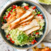 vooraanzicht van een diep bord met parelcouscous met veel groentjes, met schijfjes gebakken kip erop en een lepeltje guacamole