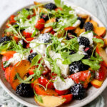 vooraanzicht van een grote schotel met een salade van perzik, kerstomaat en braambessen met balsamicovinaigrette. Afgewerkt met lekker veel burrata en rucola.