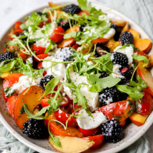 vooraanzicht van een grote schotel met een salade van perzik, kerstomaat en braambessen met balsamicovinaigrette. Afgewerkt met lekker veel burrata en rucola.