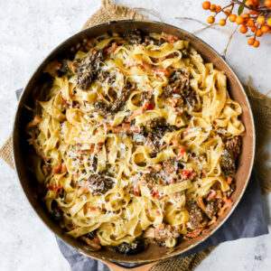 bovenaanzicht van een grote pan met daarin een romige tagliatelle met boschampignons en mascarpone, met spekblokjes, rode peper, bieslook en pecorino.